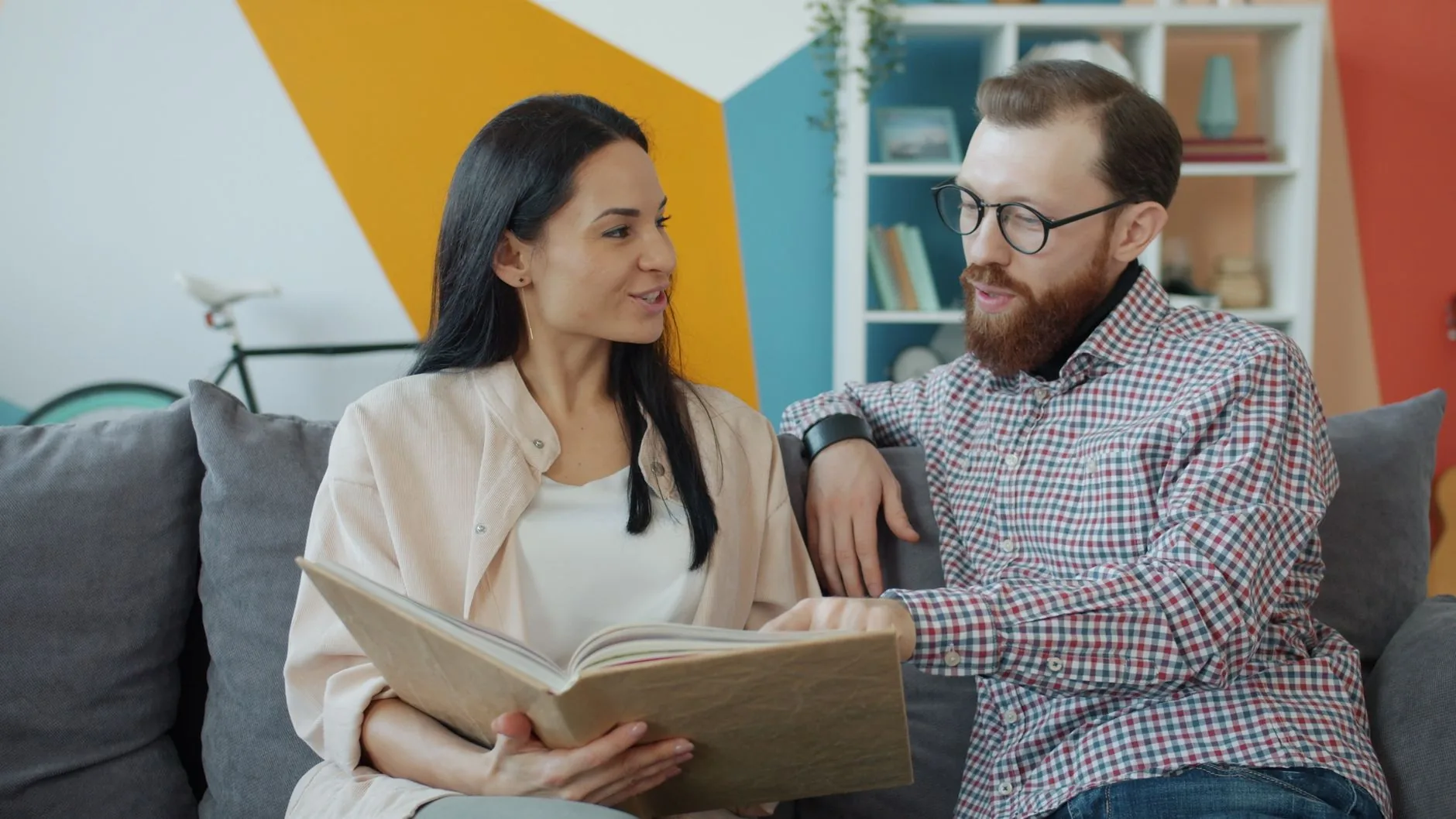 Deux personnes consultent ensemble un livre ouvert, assis sur un canapé dans un intérieur moderne et coloré