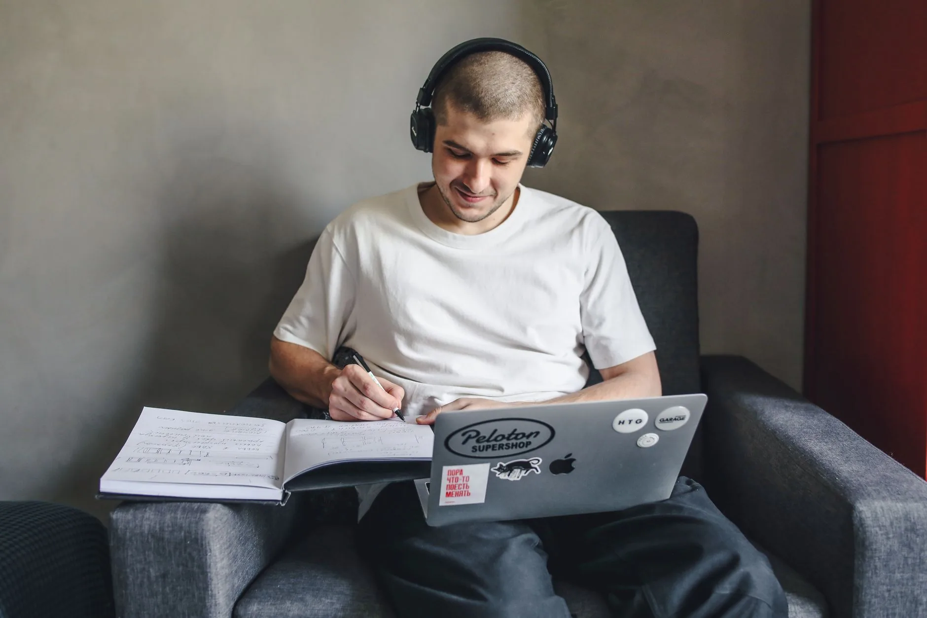 Personne portant des écouteurs, écrivant dans un carnet tout en consultant un ordinateur portable, assise dans un fauteuil