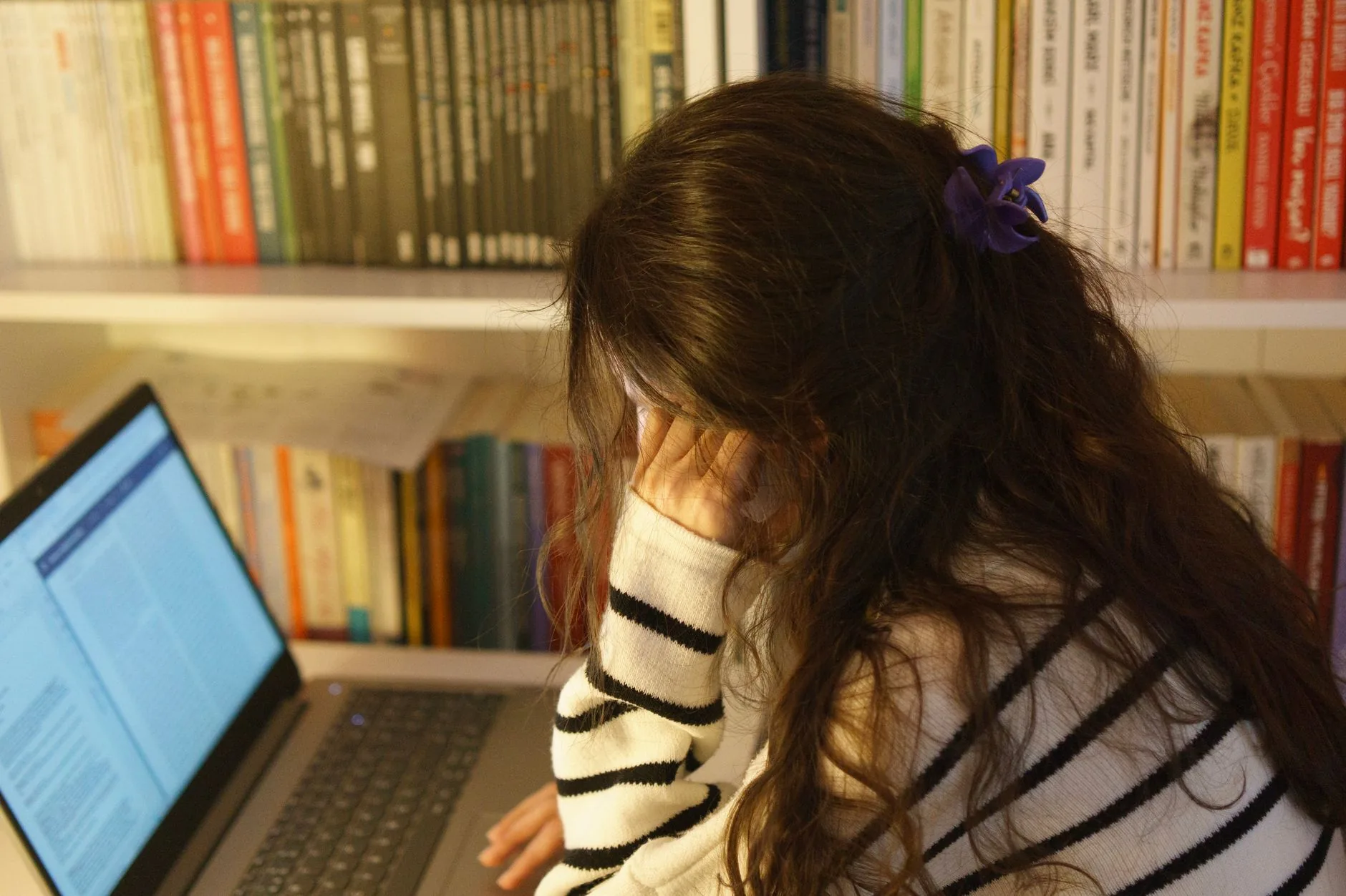 Personne assise devant un ordinateur portable, dos à la caméra, devant une bibliothèque remplie de livres