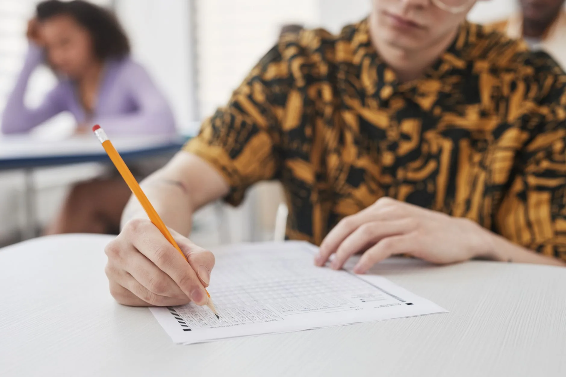 Personne remplissant un formulaire écrit au crayon, assise à une table avec d'autres personnes en arrière-plan