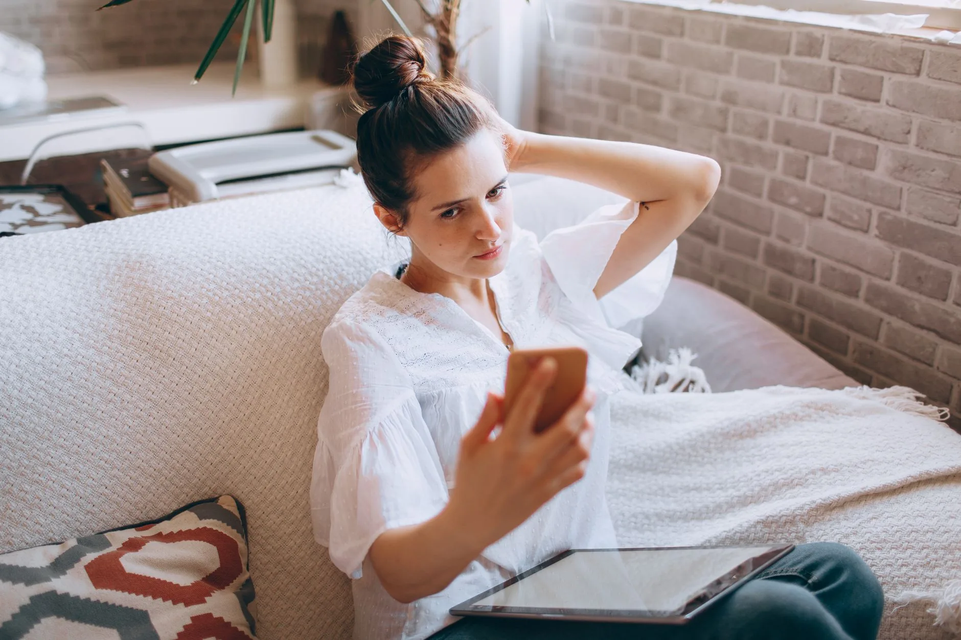 Une femme consulte son smartphone sur un canapé, une tablette posée sur ses genoux